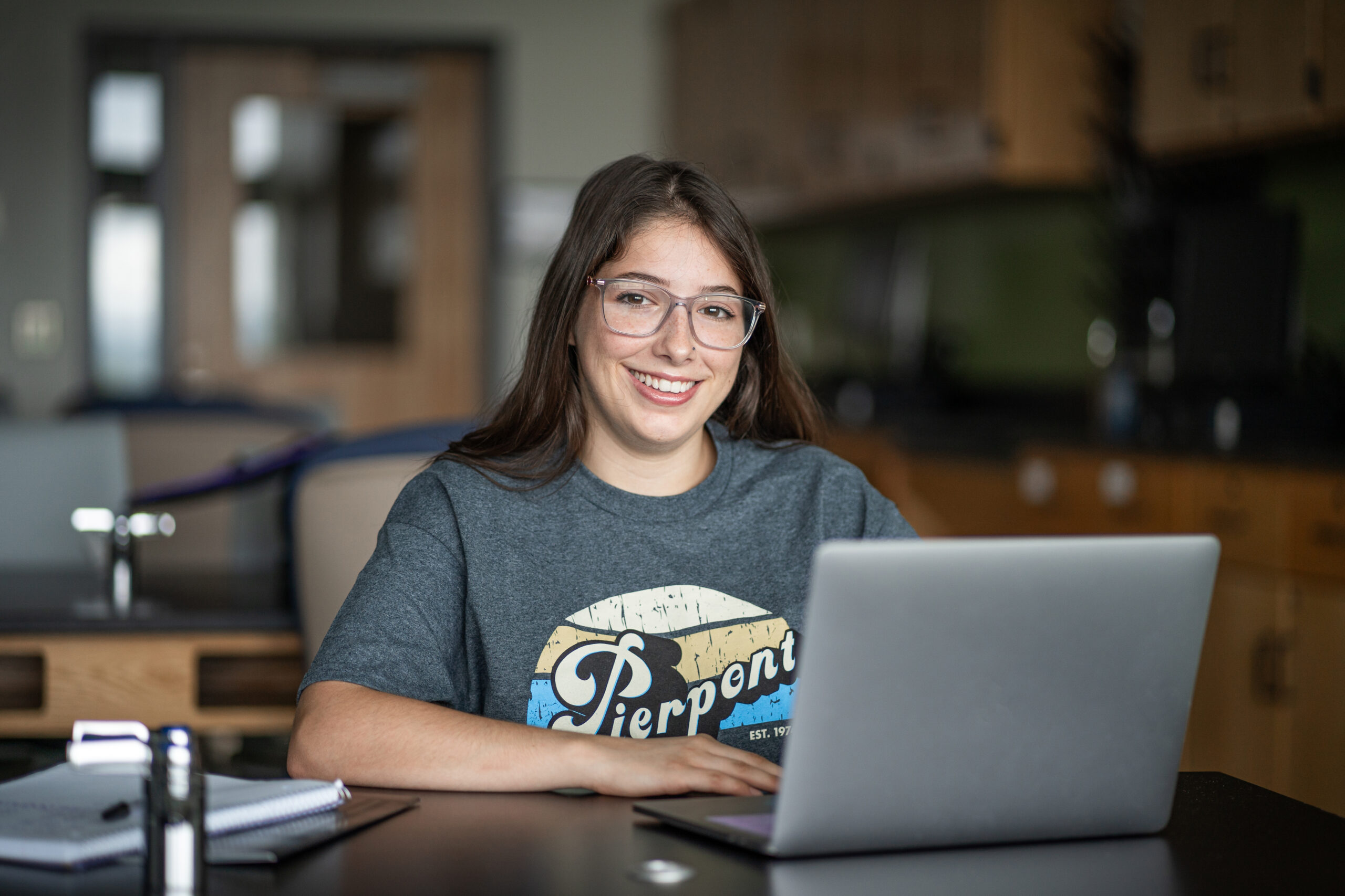 A student smiles in a classroom setting while sitting in front of a laptop.