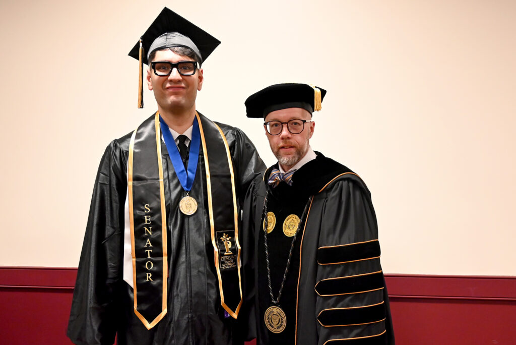 Graduate in cap and gown stands next to a Pierpont Community & Technical College official in academic regalia, both facing the camera during commencement.