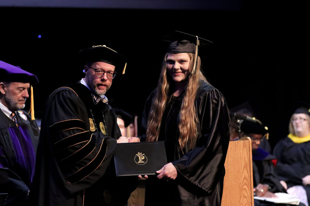 Pierpont Community & Technical College official hands a diploma to a graduate during the commencement ceremony, with a podium and faculty visible in the background.