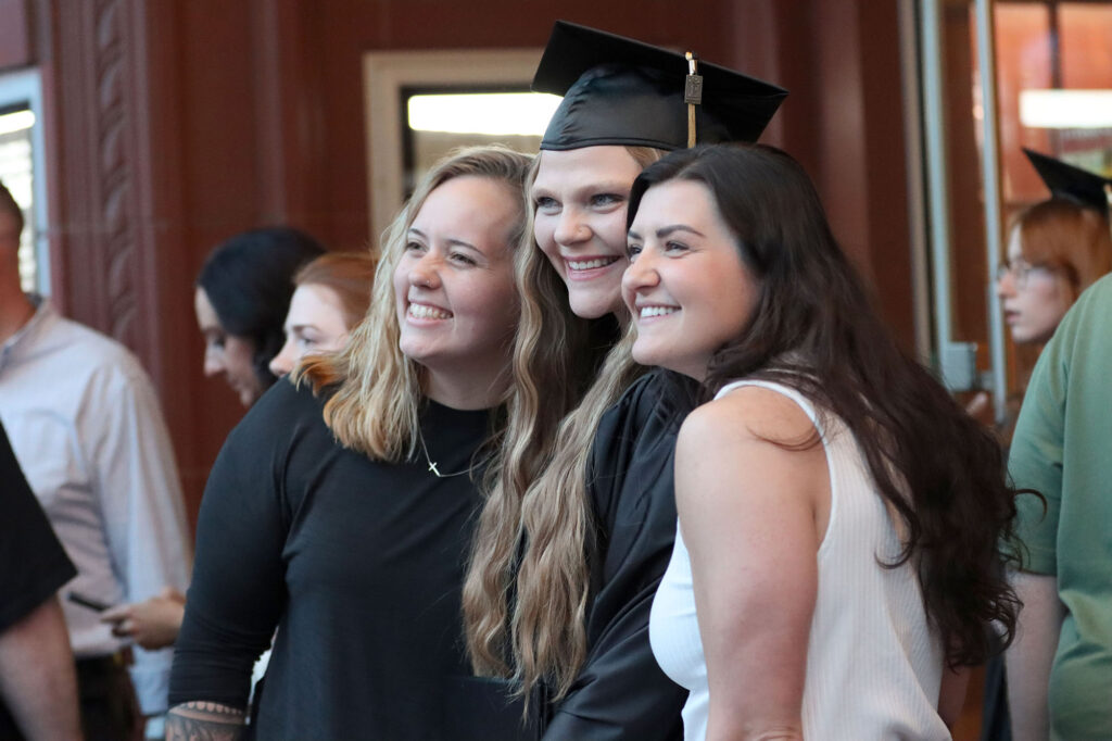 Graduate in cap and gown stands with two others while posing for a photo in a lobby area after a commencement ceremony.