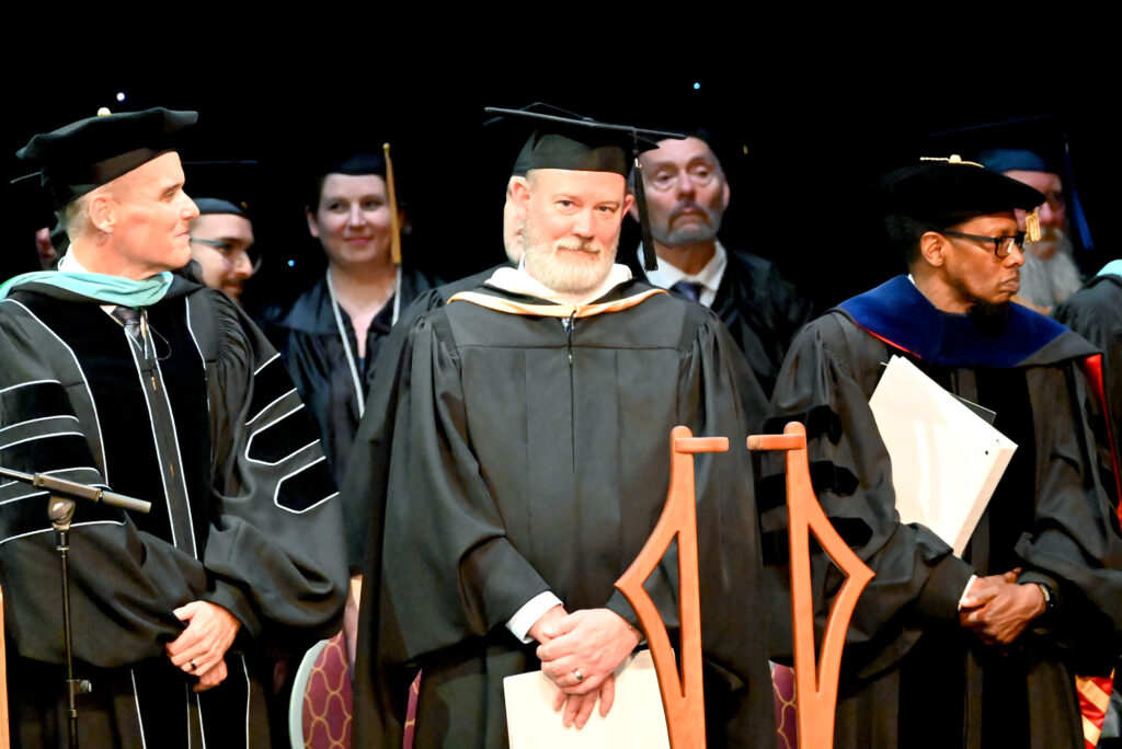 Commencement faculty and leadership in academic regalia stand on stage near a ceremonial mace during a commencement ceremony.