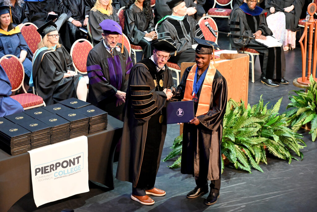 College president shakes hands with a graduate holding a diploma cover on stage during a commencement ceremony, with faculty seated behind.