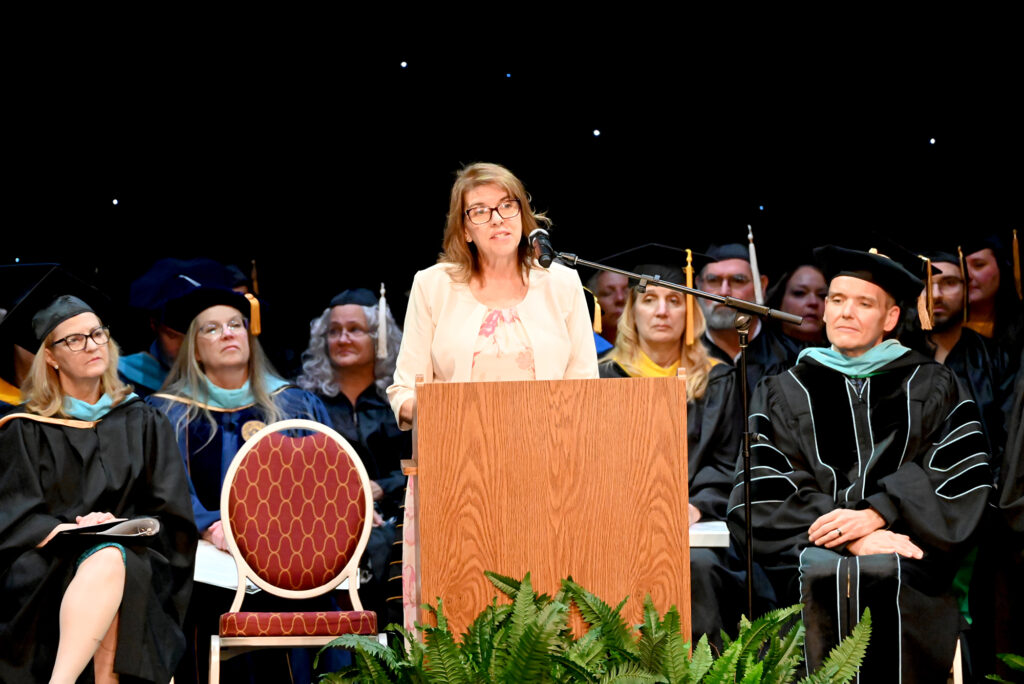 A speaker stands at a podium addressing the audience during a college commencement ceremony, with faculty seated behind on stage.