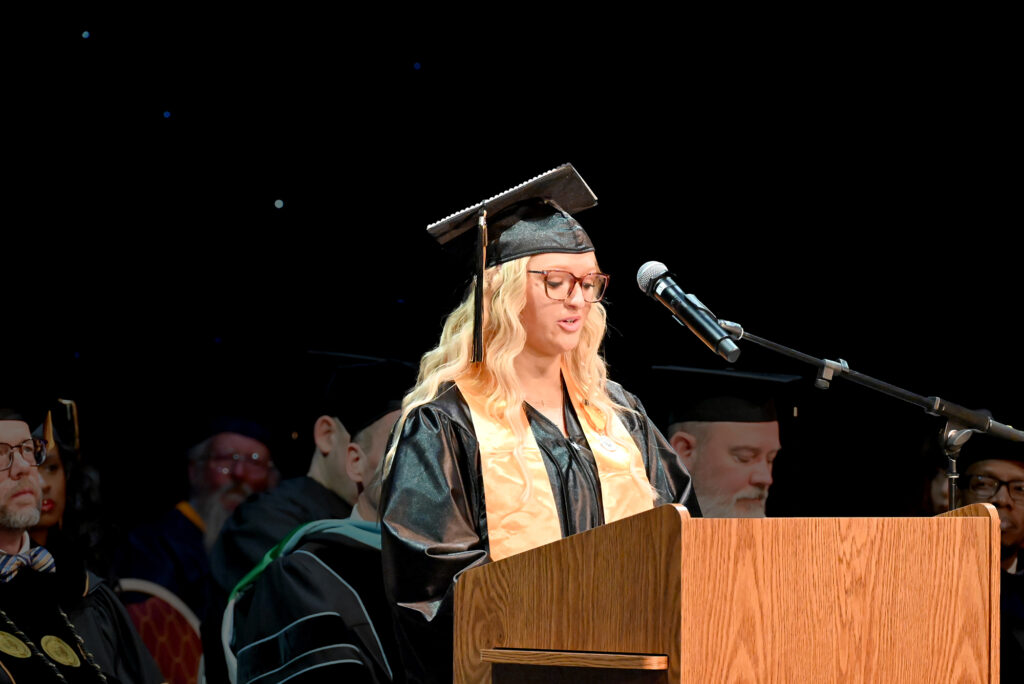 A graduate in cap and gown speaks at a podium during a commencement ceremony, with faculty visible in the background.