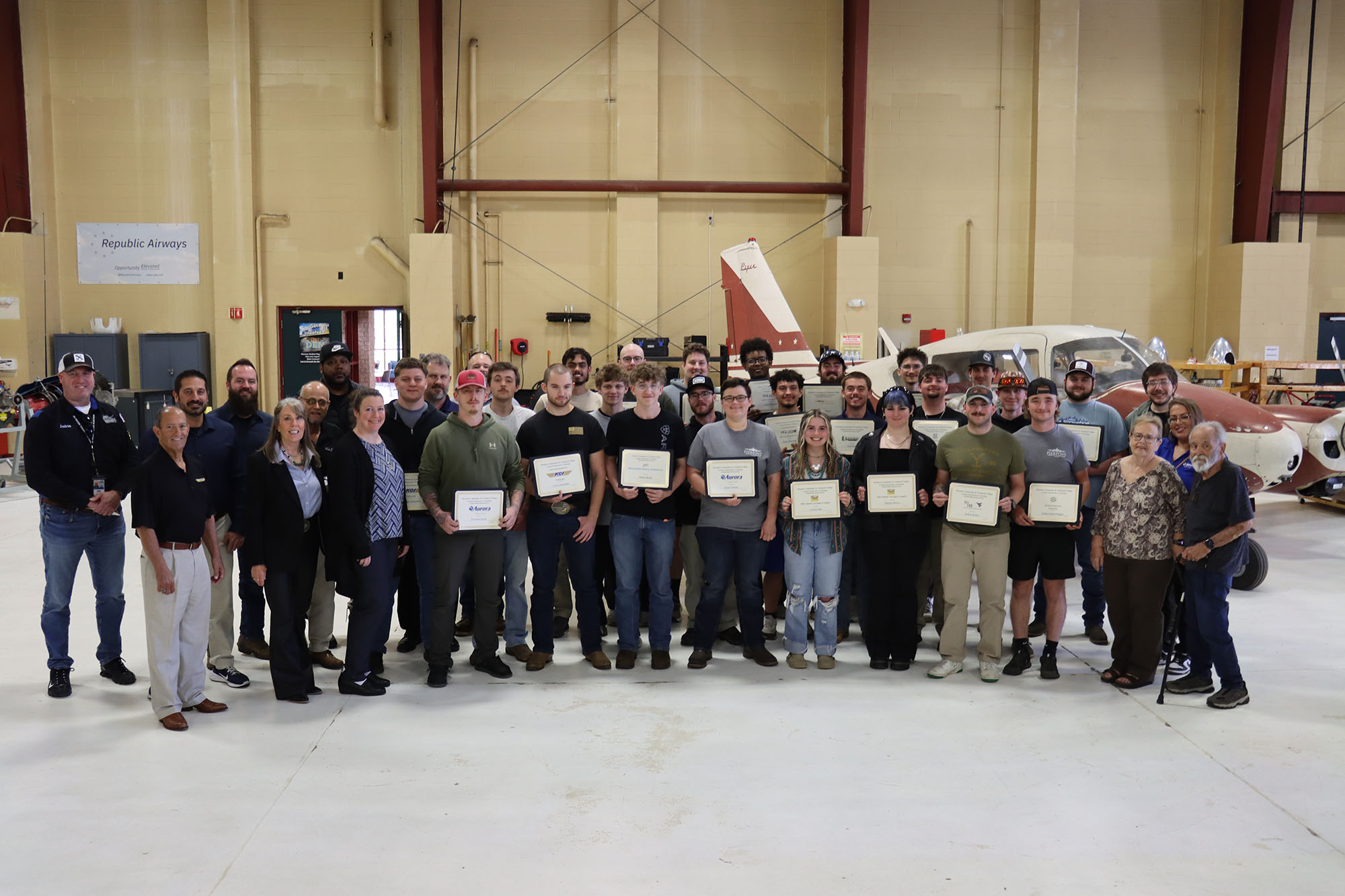 A large group of aviation maintenance technology students and industry representatives stand together in a hangar, with several individuals holding framed scholarship certificates in front of aircraft and equipment.