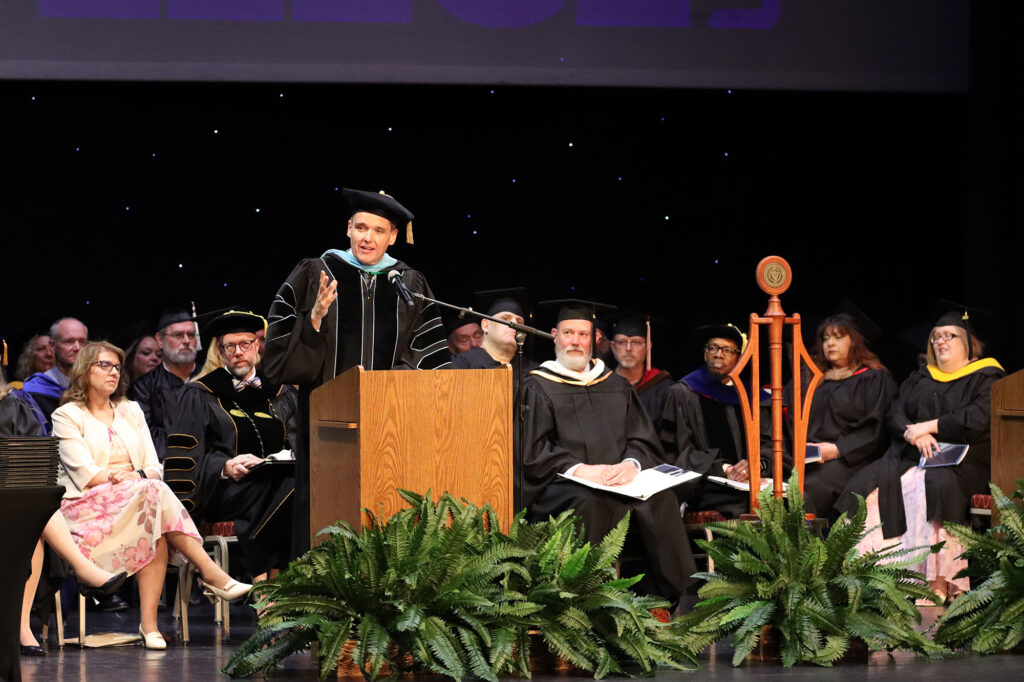 A state official speaks at a podium during a commencement ceremony, with faculty in academic regalia seated on stage beside a ceremonial mace.