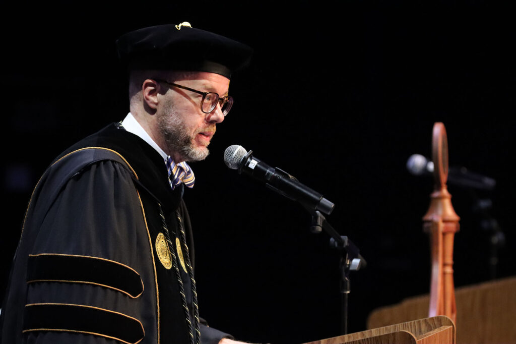The college president in academic regalia speaks into a microphone at a podium during a commencement ceremony, with stage elements visible in the background.