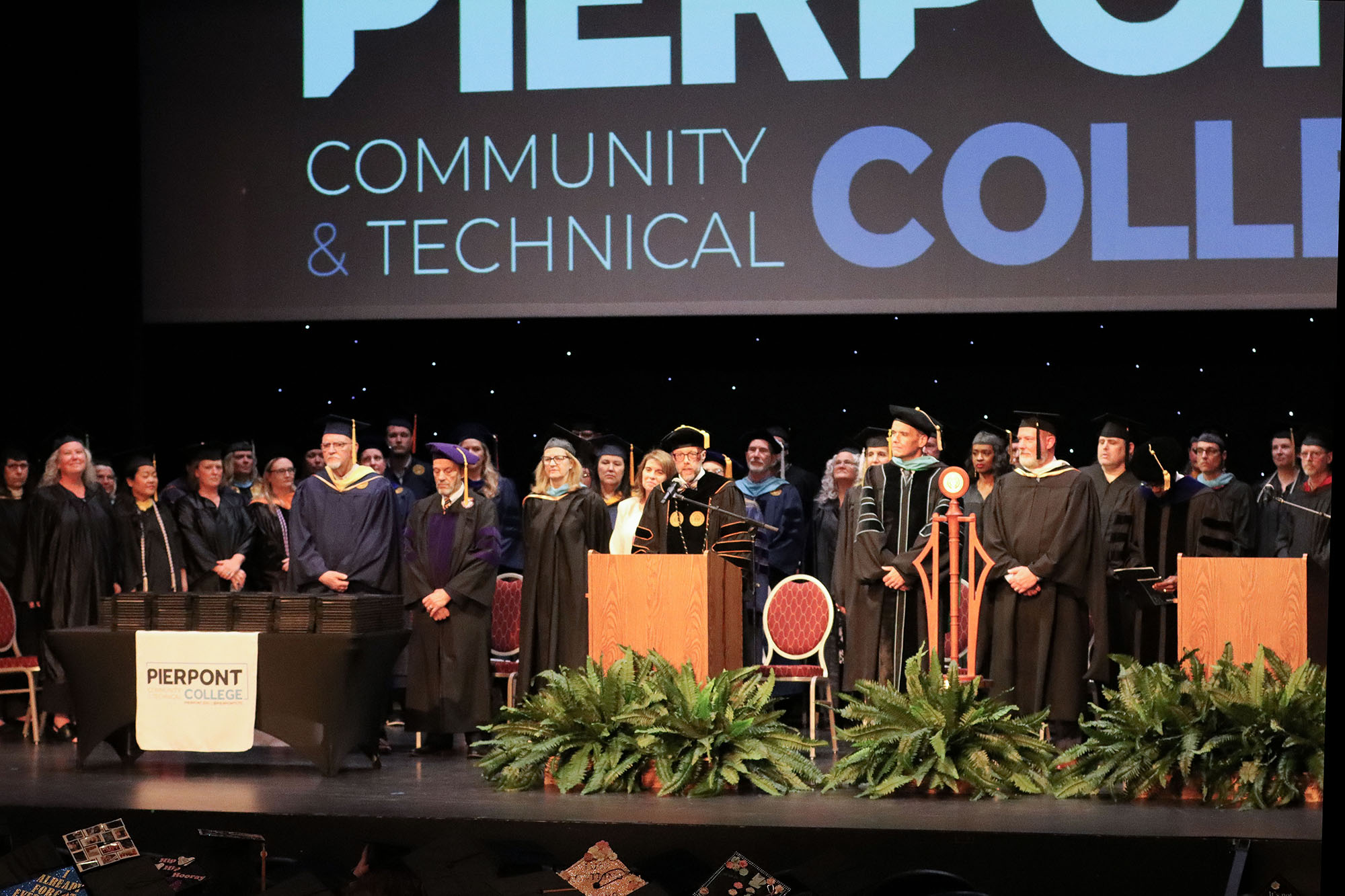 Wide view of a commencement ceremony stage with faculty in academic regalia standing behind a podium, a ceremonial mace displayed nearby, and stacks of diploma covers at the side.